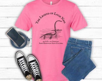 May include: Pink t-shirt with a black graphic of the Loch Ness Monster and the text "The Legend of Loch Ness" and "565 A.D. - 1st Sighting Irish Missionary Saint Columba".
