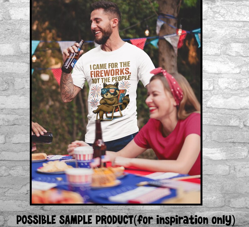 Op de afbeelding: Wit T-shirt met de tekst "I CAME FOR THE FIREWORKS, NOT THE PEOPLE" en een wasbeer met zonnebril. De wasbeer zit in een strandstoel met vuurwerk op de achtergrond. Het shirt wordt door een persoon gedragen.