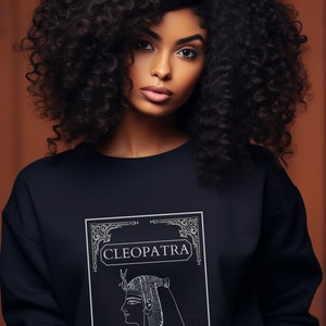 Può includere: Una felpa blu navy con una grafica bianca di Cleopatra, con il testo "CLEOPATRA" e "THE ULTIMATE FEMME FATALE AND POLITICAL BAD ASS". La modella ha i capelli ricci scuri.