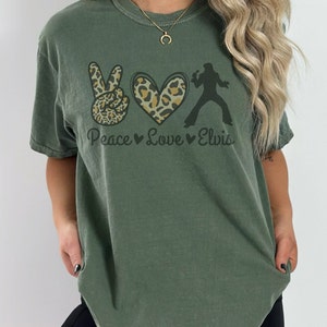 K&ouml;nnte beinhalten: Ein gr&uuml;nes T-Shirt mit einem Peace-Zeichen, einem Herzen und einer Silhouette von Elvis Presley. Das Peace-Zeichen und das Herz sind mit einem Leopardenmuster verziert. Der Text "Peace Love Elvis" ist unter der Grafik gedruckt.
