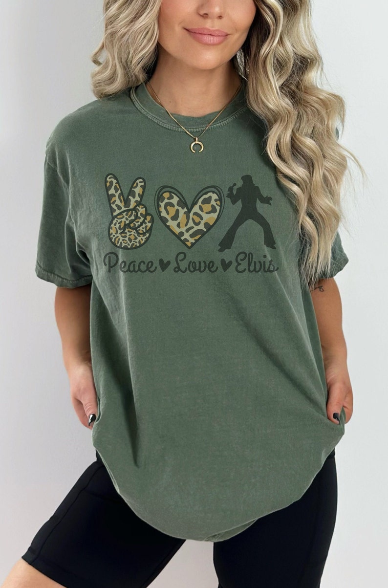 K&ouml;nnte beinhalten: Ein gr&uuml;nes T-Shirt mit einem Peace-Zeichen, einem Herzen und einer Silhouette von Elvis Presley. Das Peace-Zeichen und das Herz sind mit einem Leopardenmuster verziert. Der Text "Peace Love Elvis" ist unter der Grafik gedruckt.