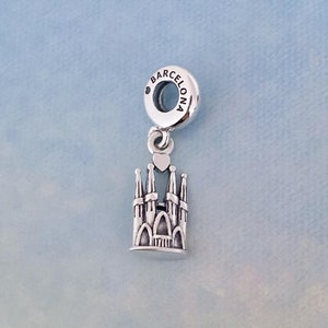 May include: Silver charm featuring a detailed depiction of the Sagrada Familia basilica in Barcelona, Spain. The charm has a small heart at the top and a round bead with the word "BARCELONA" engraved on it.