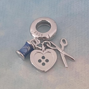 Puede incluir: Un charm plateado con elementos de costura. Presenta un carrete de hilo azul, un botón en forma de corazón y unas tijeras. El charm está detallado con la inscripción "S925" y está diseñado para la fabricación de joyas.