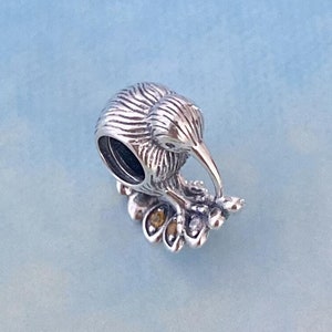 May include: Silver charm featuring a detailed kiwi bird perched on a branch with small leaves. The charm is designed to be used on a bracelet or necklace.