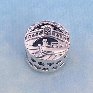 May include: Silver charm featuring a detailed depiction of a gondola under a bridge with a heart-shaped design. The charm is adorned with clear cubic zirconia stones.
