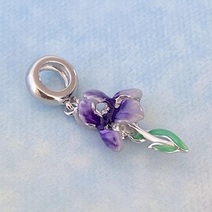 May include: A silver charm featuring a purple flower with green leaves. The charm has a loop for attaching to a bracelet or necklace.