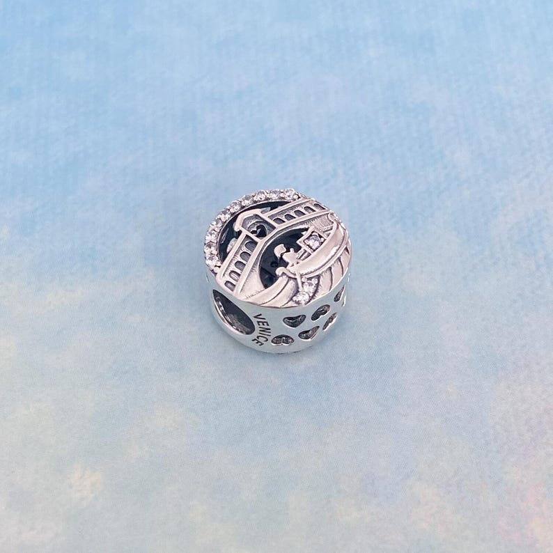May include: Silver charm featuring a detailed depiction of the Rialto Bridge in Venice, Italy. The charm is adorned with clear cubic zirconia stones and the word "VENICE" is engraved on the side.