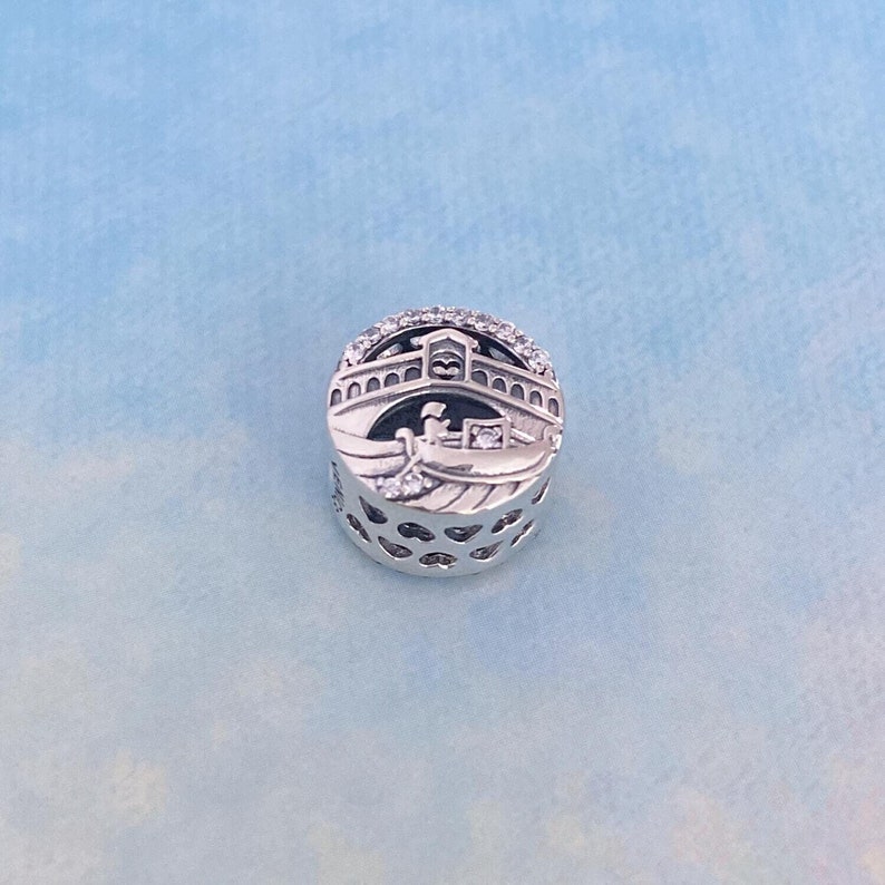 May include: Silver charm featuring a detailed depiction of a gondola under a bridge with a heart-shaped design. The charm is adorned with clear cubic zirconia stones.