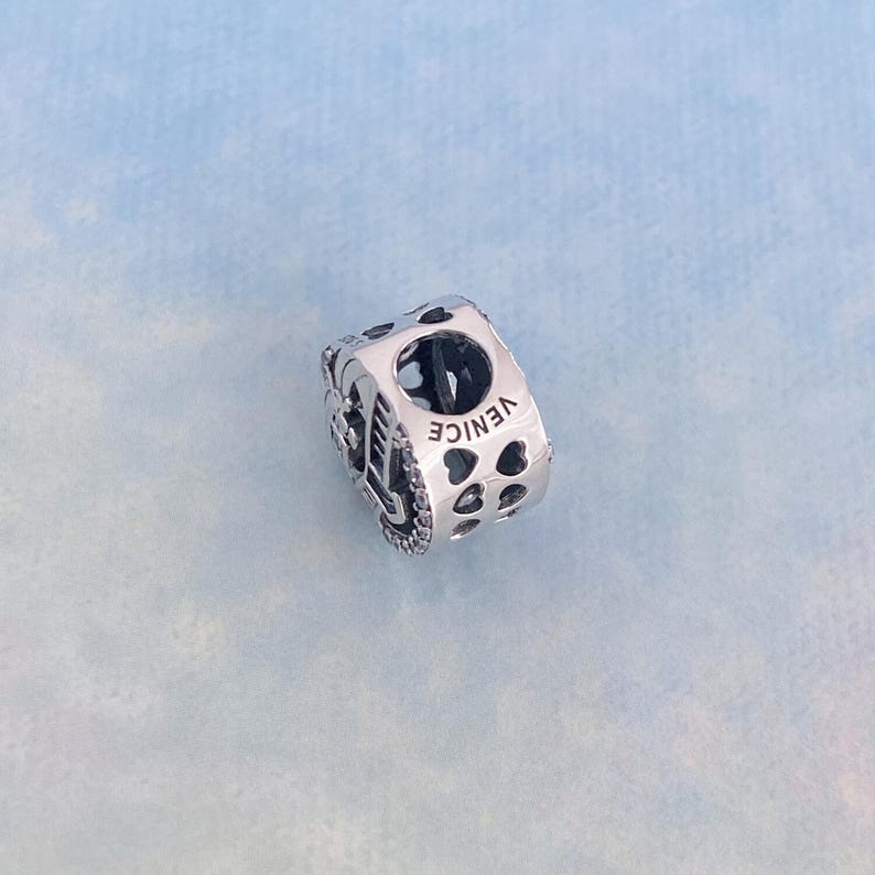 May include: Silver charm with a detailed design of a gondola and the word "VENICE" engraved on the side. The charm is decorated with small heart cutouts.