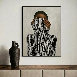 Op de afbeelding: Een ingelijste kunstdruk met een persoon met het gezicht gedeeltelijk bedekt door een zwart-wit patroon kledingstuk. Een donkerbruine vaas staat op een houten oppervlak.