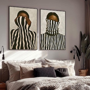 Op de afbeelding: Twee ingelijste wandkunstwerken. Elk toont een persoon met een zwart-wit gestreepte coltrui. Eén portret toont de persoon die zijn ogen bedekt, de andere met gesloten ogen. De kunst wordt boven een bed tentoongesteld.