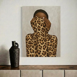 Op de afbeelding: Een canvas print met een persoon gehuld in een luipaardprint stof, handen bedekken het gezicht. Het kunstwerk heeft een neutrale achtergrond en wordt weergegeven boven een houten oppervlak. Een donkere, getextureerde vaas staat links.