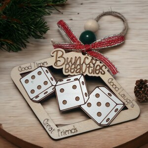 May include: Wooden Christmas ornament shaped like a square frame with three dice inside. The ornament is decorated with the text "Bunco Beauties", "Good Times", "Good Laughs", and "Great Friends".