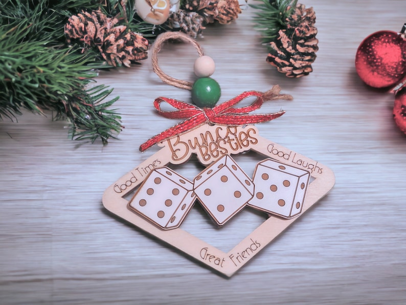 May include: Wooden Christmas ornament shaped like a dice frame with three dice inside. The ornament is decorated with a red bow and the words "Bunch besties", "Good Times", "Good Laughs", "Great Friends".