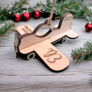 May include: Wooden aeroplane ornament with the text "Merry Christmas" and "2023" on the wings and the name "Eli" on the body. The ornament is decorated with red and green Christmas baubles.