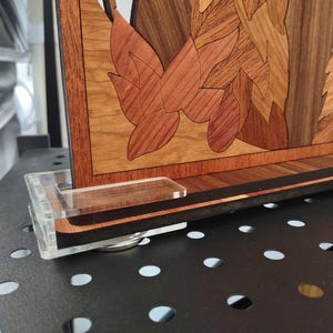 Può includere: Un'opera d'arte murale in legno con un design floreale, che presenta dettagli intricati e varie tonalità di marrone. L'opera è esposta su un supporto in acrilico trasparente.