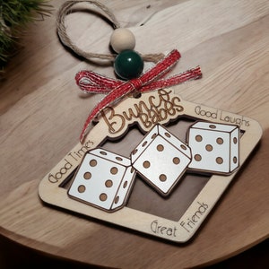 May include: Wooden Christmas ornament shaped like a dice with the words "Bunco Babes" and "Good Times, Good Laughs, Great Friends" engraved on it. The ornament is decorated with a red and silver ribbon.