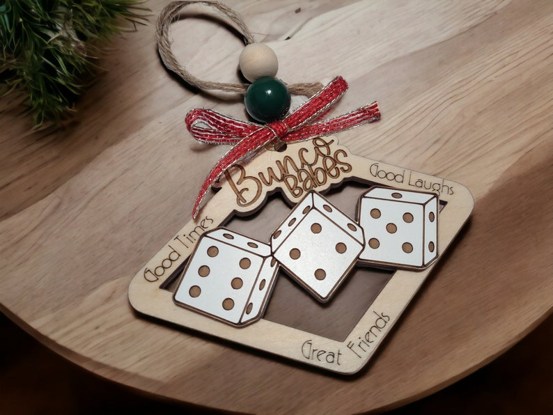 May include: Wooden Christmas ornament shaped like a dice with the words "Bunco Babes" and "Good Times, Good Laughs, Great Friends" engraved on it. The ornament is decorated with a red and silver ribbon.