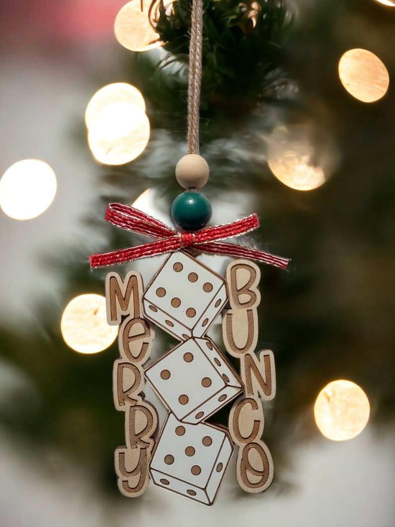 May include: A wooden Christmas bauble with the words "Merry Bunco" and three dice. The bauble is decorated with a red and white ribbon and a green and wooden bead.