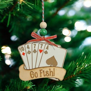May include: A wooden Christmas bauble shaped like a hand of playing cards with the text "Go Fish!" on a banner. The cards are a 10 of diamonds, 4 of spades, 7 of clubs, 7 of hearts, and an ace of spades.