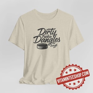 May include: A light beige t-shirt with the text "Dirty Fuckin' Dangles boys" in a stylized font, accompanied by a hockey puck graphic. The shirt has short sleeves and a crew neck.