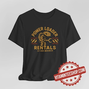 May include: Black t-shirt with a gold and brown graphic design. The design features the text "POWER LOADER RENTALS LV-426 BRANCH" with an illustration of a power loader machine. A red and white seal is in the bottom right corner.