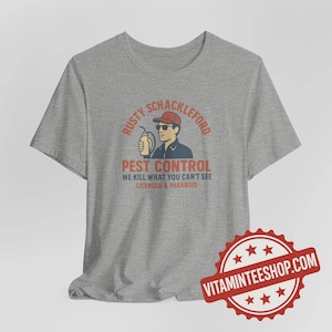 May include: A heather gray t-shirt featuring a graphic for "Rusty Shackleford Pest Control." The design includes a cartoon man in a cap and sunglasses holding a sprayer, with the text "We kill what you can't see. Licensed & Paranoid."