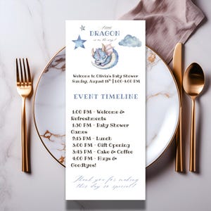 May include: A baby shower invitation with a watercolor dragon illustration and the text "A Dragon is on the way!". The invitation includes the event details and timeline, set on a marble plate with gold cutlery.