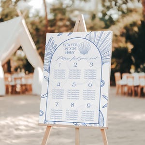 Puede incluir: Un letrero blanco sobre un caballete de madera con el texto "SEA YOU SOON BABY" y "Please find your seat." El letrero tiene un diseño azul de conchas marinas y botánico y una tabla de asientos numerada.