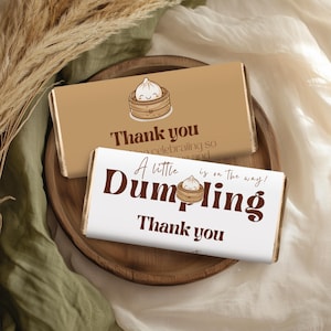 May include: Two chocolate bar wrappers with a white background and brown text. The top wrapper has a brown background with a cartoon dumpling in a steamer basket. The text reads "Thank you for celebrating so much love and joy". The bottom wrapper has the text "A little Dumpling is on the way! Thank you".