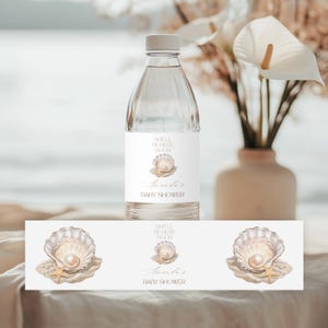 May include: A clear water bottle with a white label featuring a seashell design and the text "Shell Be Here Soon" and "Amanda's Baby Shower". A matching label is below the bottle. A vase of flowers is in the background.