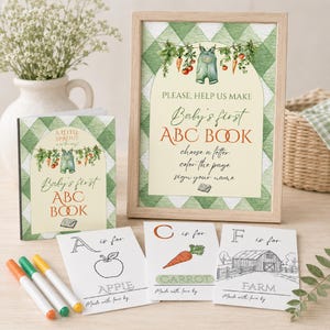 May include: A framed sign and a book titled Baby's first ABC BOOK with a green gingham background and garden illustrations. Three coloring cards labeled A, C, and F with apple, carrot, and farm drawings are displayed with three colored markers.