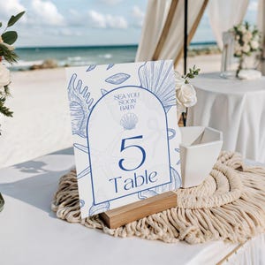 May include: A white table number card with blue seashell illustrations and the text "SEA YOU SOON BABY" and "5 Table". The card is displayed on a wooden base, with a macrame table runner and beach scene in the background.