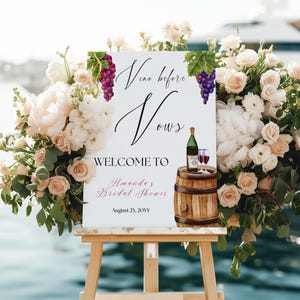Puede incluir: Un cartel blanco con el texto "Vino before Vows" en caligrafía negra. El cartel también dice "Welcome to Amanda's Bridal Shower" en negro con "Amanda's" en rosa. La fecha "August 25, 20YY" también está incluida en negro. El cartel está decorado con uvas de acuarela y un barril con una botella de vino y dos vasos.