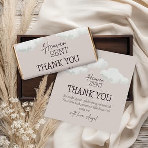May include: A light beige chocolate bar and card set with the words "Heaven Sent Thank You" in elegant script. The card includes a thank you message and is decorated with cloud illustrations. The set is presented on a wooden tray.