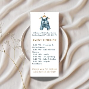 Pode incluir: Um convite para chá de bebê com uma ilustração em aquarela de macacão jeans em um cabide de madeira. O convite inclui o texto "Blue Jean Baby", detalhes do evento e um cronograma de atividades. O convite está em um fundo branco.