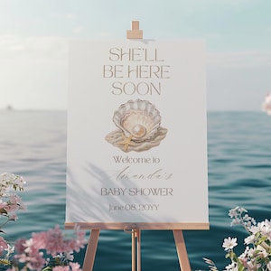 Könnte beinhalten: Ein weißes rechteckiges Schild auf einer Holzstaffelei mit dem Text SHE'LL BE HERE SOON, Welcome to Amanda's BABY SHOWER und June 08, 20YY. In der Mitte befindet sich eine Aquarellillustration einer offenen Muschel mit einer Perle und einem Seestern.