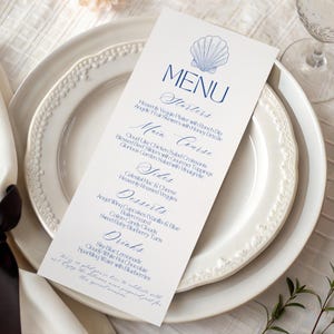 Pode incluir: Um cartão de menu formal com um gráfico de concha na parte superior, colocado em um prato branco. O menu é impresso em script azul marinho e lista entradas, pratos principais, acompanhamentos, sobremesas e bebidas. O menu é para uma ocasião especial.