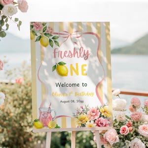 Könnte beinhalten: Ein Willkommensschild für einen ersten Geburtstag mit dem Text "Freshly ONE" und einer Zitronenillustration. Das Schild ist mit rosa und gelben Blumen, Zitronen und einem rosa Band verziert. Das Schild steht auf einer Staffelei.