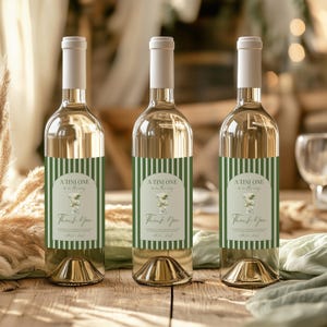 May include: Three wine bottles with white labels featuring green striped borders and the text "A TINI ONE" and "Thank You". The bottles have a clear glass body, white cap, and gold base, set on a wooden surface.