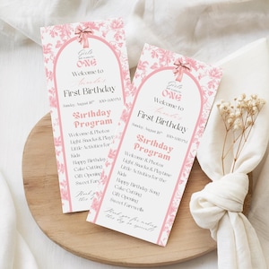 May include: Two pink and white birthday program cards with floral patterns and text. The cards read "Girls just wanna be ONE" and list the birthday program schedule. The cards are on a wooden tray.