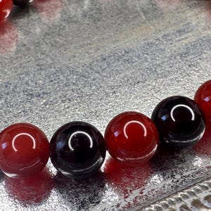 May include: A close-up of a bracelet made of alternating red and black beads. The beads are round and smooth, and they are strung together on a clear elastic cord.