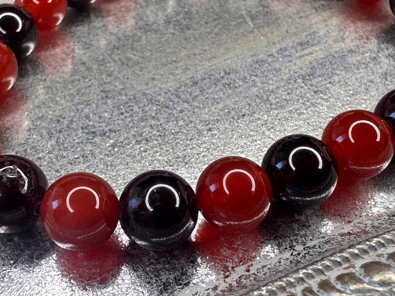 May include: A close-up of a bracelet made of alternating red and black beads. The beads are round and smooth, and they are strung together on a clear elastic cord.