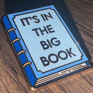 Pode incluir: Um broche de esmalte azul em forma de livro com o texto "It's in the Big Book" em letras pretas.