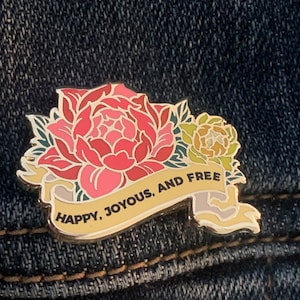 May include: A gold-toned enamel pin featuring a pink and red flower with green leaves and a yellow banner that reads "Happy, Joyous, and Free".