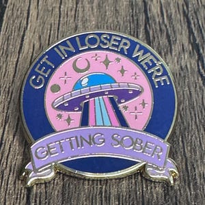 Può includere: Uno spillo in smalto dorato con un'astronave aliena blu e viola che vola su uno sfondo rosa con stelle e una luna crescente. Il testo "Get in Loser We're Getting Sober" è scritto attorno all'astronave in un carattere dorato.