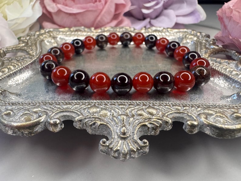 May include: A red and black gemstone bracelet with alternating beads. The bracelet is on a silver tray.
