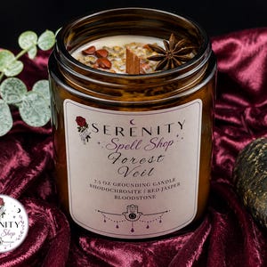 May include: A 7.5 oz amber glass candle jar with a wooden wick, topped with red jasper, rhodochrosite, and a star anise. The label reads Serenity Spell Shop Forest Veil, 7.5 oz Grounding Candle, Rhodochrosite, Red Jasper, Bloodstone with a hamsa icon.
