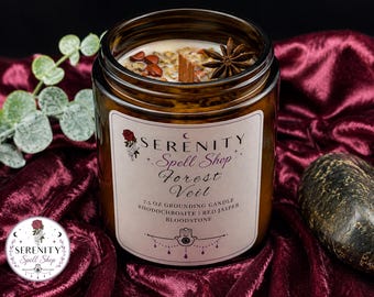 May include: A 7.5 oz amber glass candle jar with a wooden wick, topped with red jasper, rhodochrosite, and a star anise. The label reads Serenity Spell Shop Forest Veil, 7.5 oz Grounding Candle, Rhodochrosite, Red Jasper, Bloodstone with a hamsa icon.