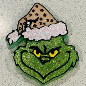 May include: Green Grinch face with a brown and white Santa hat with white glitter. The Grinch has yellow eyes and a black outline.